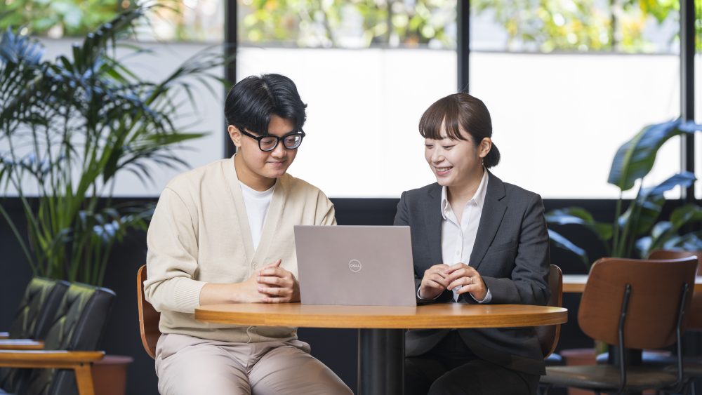 東洋言語学院の独自学習・生活支援システム『4つのM』のMy Teacher(個別伴走)による面談風景。1つのテーブルで先生と留学生がパソコン画面を笑顔で一緒に確認しながら、学習や進路の相談に乗っている様子。